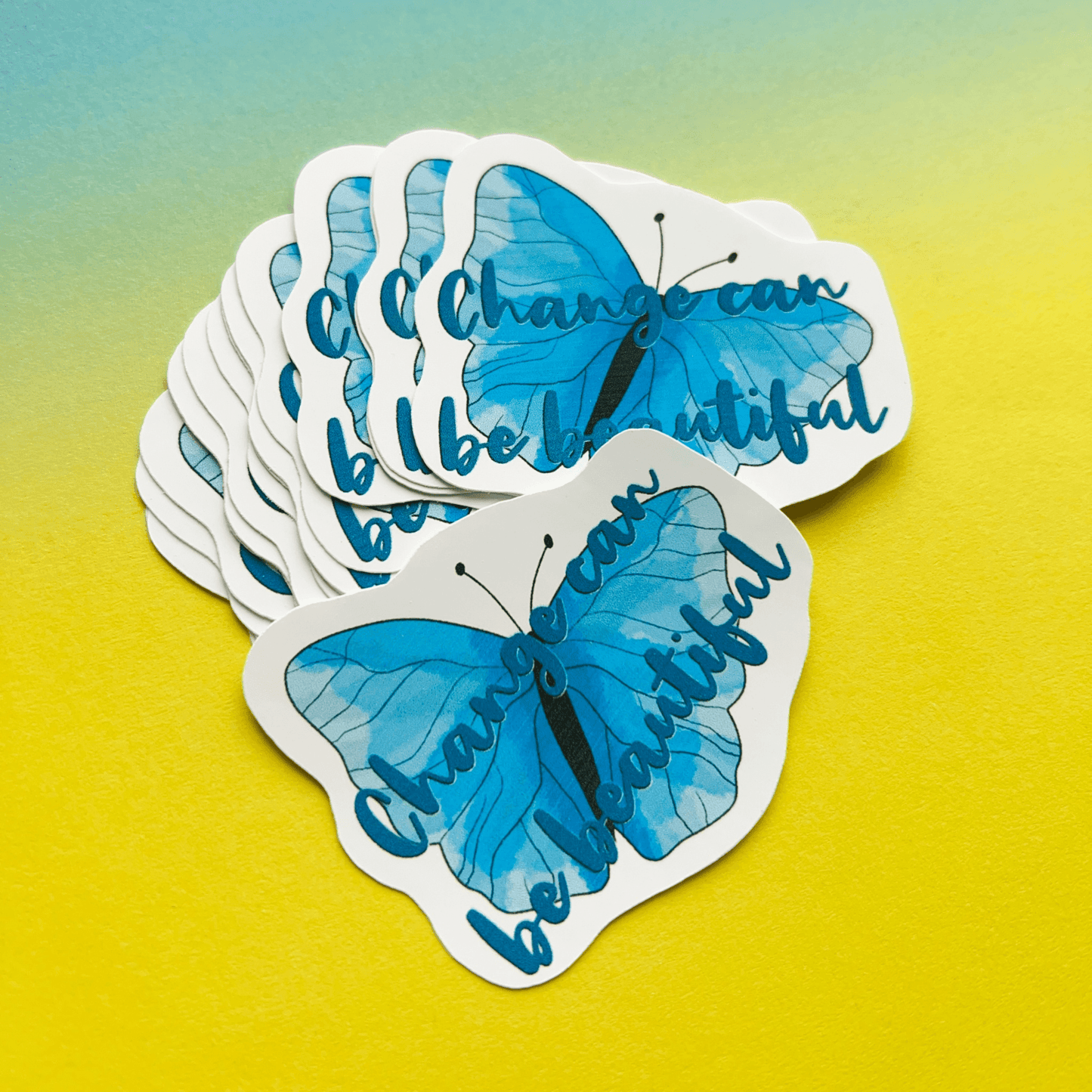 A stack of "Change can be Beautiful" Vinyl Stickers features blue butterflies and an uplifting message set against a gradient yellow to green background. These matte vinyl motivational stickers add an inspiring touch wherever they are placed.