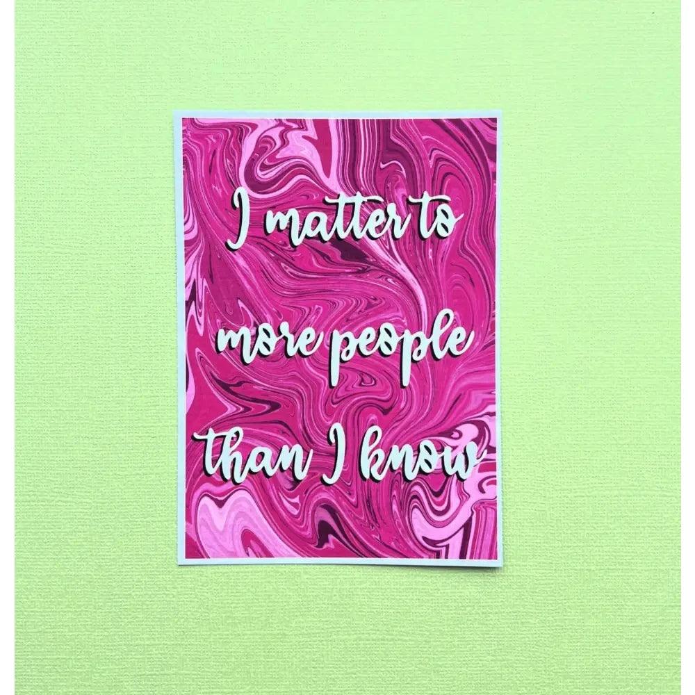 An Affirmation Postcard featuring a pink and white marbled background with the inspirational quote "I matter to more people than I know" beautifully scripted in elegant white cursive letters. The postcard is elegantly displayed on a light green textured surface.