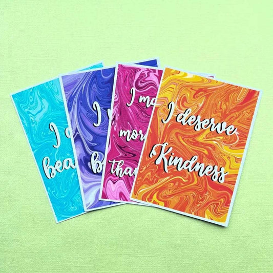 Four Affirmation Postcards with vibrant, marbled backgrounds rest on a light green surface. Each card is adorned with an inspirational quote in white cursive font: "I am beautiful," "I am brave," "I am more than enough," and "I deserve kindness.