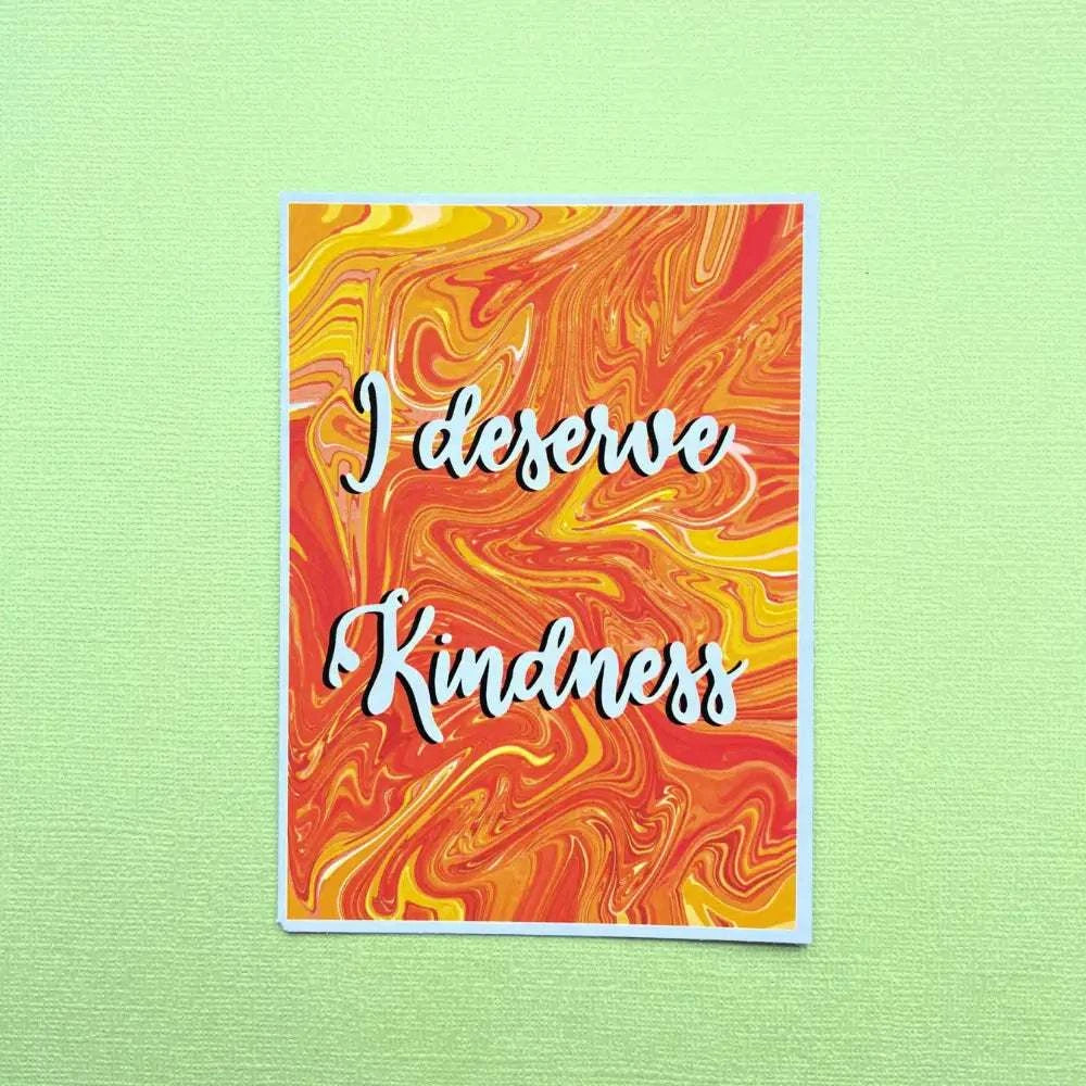 An Affirmation Postcard showcases a vibrant marbled background in shades of orange, red, and yellow, prominently displaying the inspirational quote "I deserve Kindness" in an elegant white cursive font. The card is placed on a textured, light green surface.