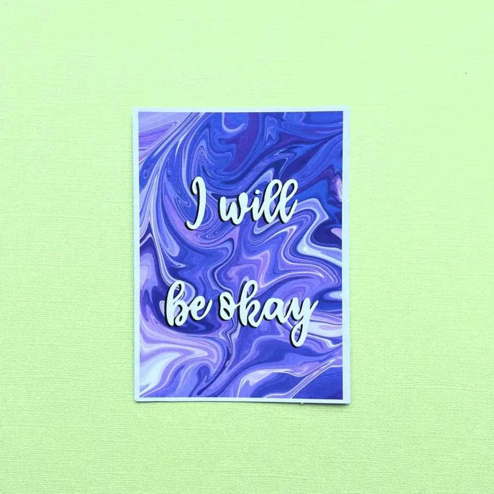 An Affirmation Postcard featuring a purple and blue marble background showcases the inspirational quote "I will be okay" in elegant white cursive letters. The postcard is set against a light green textured surface.