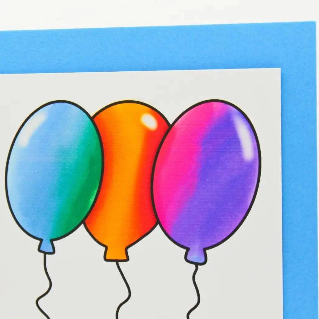 The Balloon Writing Birthday Card displays three illustrated balloons in green, orange, and purple with black strings on a white background. Placed on a light blue surface, the card features a cheerful "Happy Birthday" message. Part of the blue background is visible around the edges of the card.