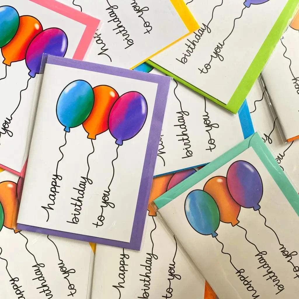 A collection of "Balloon Writing Birthday Cards," featuring vibrant images of balloons and the text "happy birthday to you" on each card, is shown scattered across the surface. These cards come in an array of colors including purple, pink, green, and yellow, and are packaged in biodegradable materials.