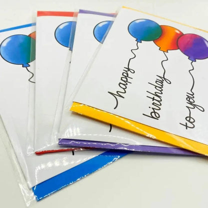 A set of Balloon Writing Birthday Cards displayed in overlapping layers, each adorned with colorful balloons and a cheerful "Happy Birthday" message on the front. The cards are bordered in vibrant colors: blue, red, purple, and yellow. They come packaged in clear biodegradable sleeves.