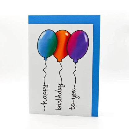 The Balloon Writing Birthday Card is a colorful birthday card featuring three balloons in blue, orange, and purple, with black strings that form the "happy birthday to you" message. The card also has a blue border and comes in biodegradable packaging.