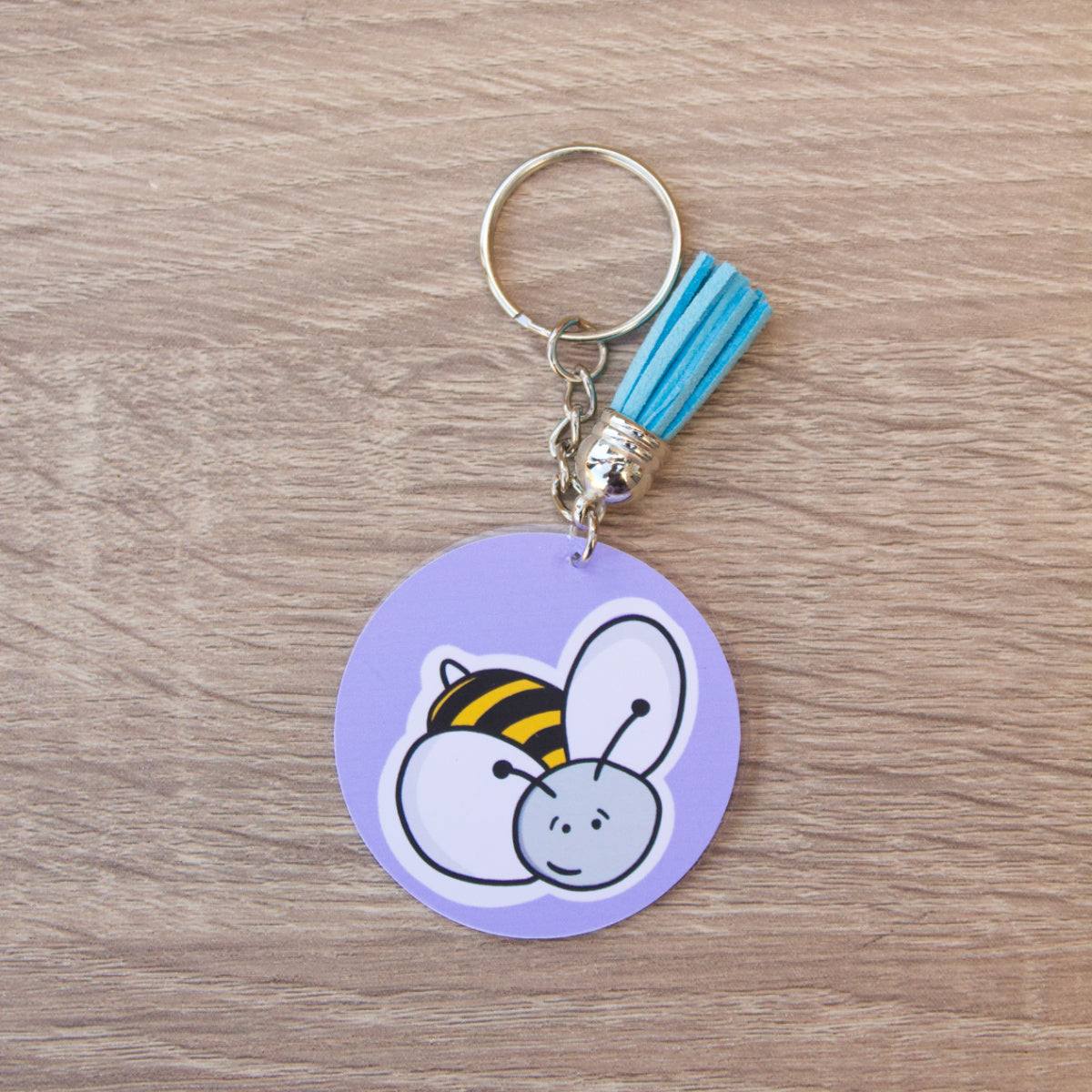 A Bumblebee Acrylic Keyring with a purple background features a cute cartoon bee with a smiling face and white wings. Attached to the keychain is a small blue tassel, all set on a wooden surface.