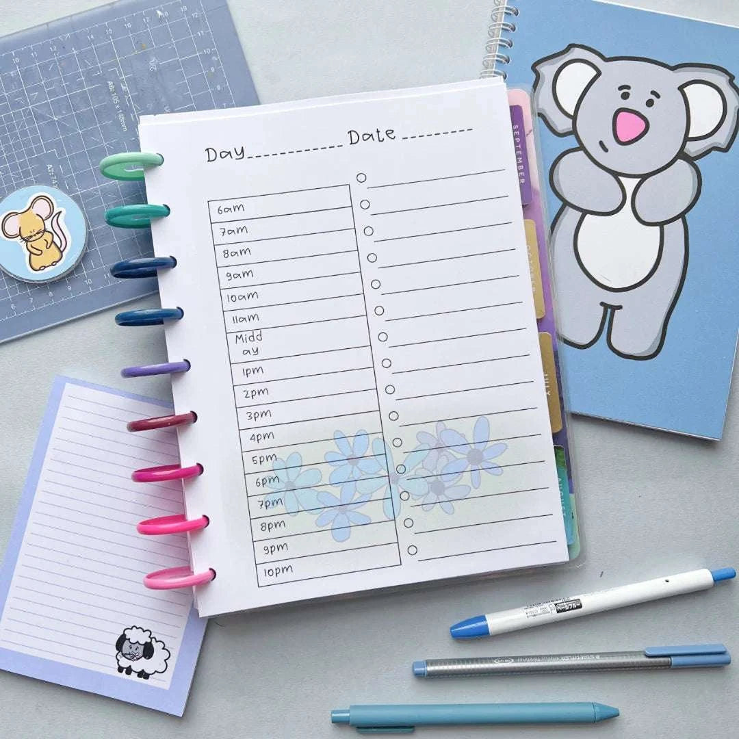 A neatly arranged workspace with a Daily Schedule and To Do List featuring hourly slots from 6am to 10pm. The planner is secured with colorful ring binders. Surrounding it are a blue notepad, a koala-themed notebook, pens, and a small cutting mat with stickers.