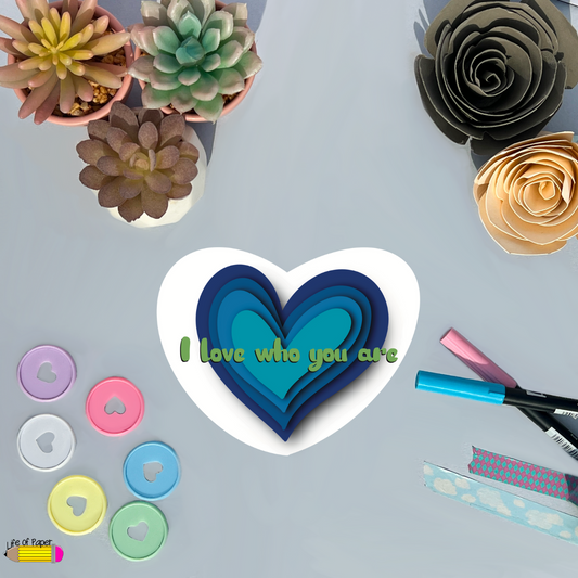 A variety of craft supplies and colorful paper flowers are arranged around a white heart with layered blue hearts inside. The "Love who you are" sticker is written in green across the heart. Markers, buttons, and a roll of water-resistant washi tape are also visible.