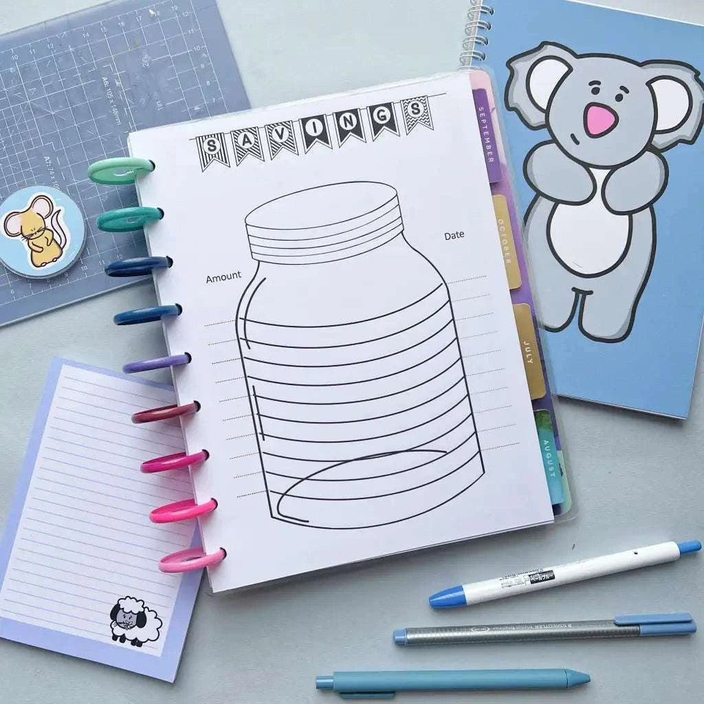 A flat lay image of a 52 Week Saving Challenge with a jar illustration at the center. Surrounding it are colorful pens, a sticker of a koala, and a notepad with a lion sticker to help keep track of your 52-week saving challenge. A notebook with a sheep illustration, pen, and pencil complete the setup.