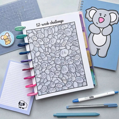 A desk with a colorful 52 Week Saving Challenge notebook. The tracker features heart shapes filled with amounts like £23 and £11, helping you save money towards your yearly savings goal. Surrounding items include pens, a blue notebook with a koala, a pad with a sheep, and a cutting mat with tools.