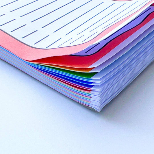 A close-up photo of a stack of multicolored lined papers displays a charming rainbow effect with edges in pink, red, green, blue, and purple. Neatly arranged against a white background, this Rainbow To Do List Desk Memo Pad creates an inviting and orderly appearance.