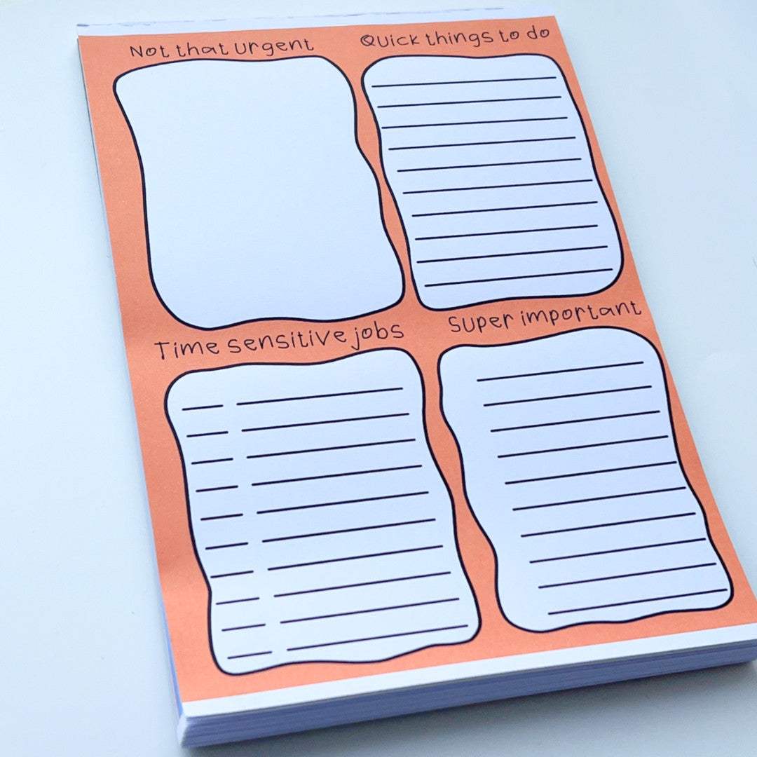 A Rainbow To Do List Desk Memo Pad with an orange border is divided into four sections: "Not that urgent," "Quick things to do," "Time-sensitive jobs," and "Super important." Each section has lines for writing tasks. The notepad is placed on a white surface.