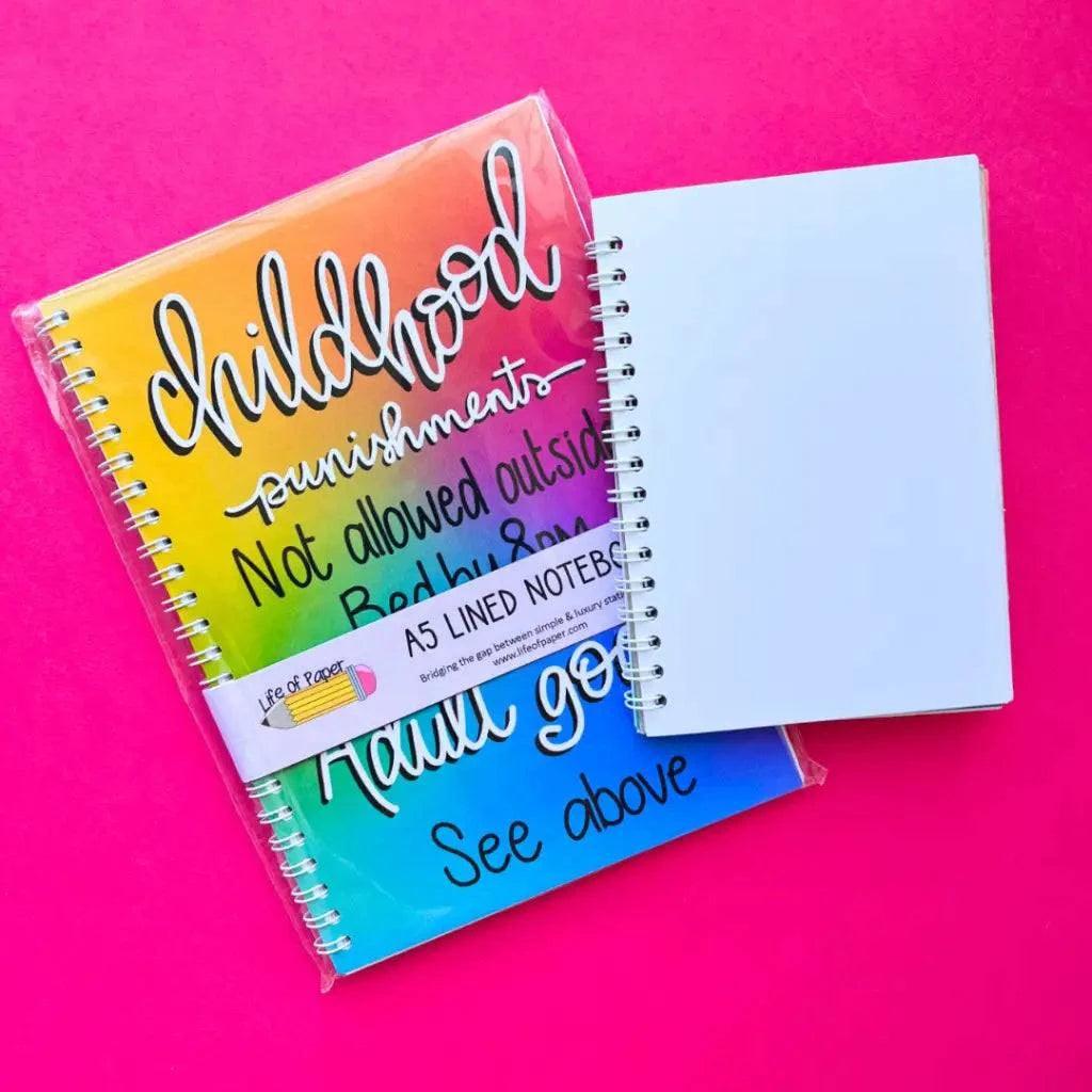 Two spiral-bound notebooks sit on a bright pink background. One, the Adulting Goals A5 Notebook, boasts a colorful ombre cover with witty text contrasting childhood punishments and adult aspirations. The other is a plain white A5 lined notebook, featuring durable 120gsm paper for a smooth writing experience.