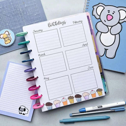 A Birthday and Celebration Calendar tracking birthdays from January to June, illustrated with cupcakes, lies open. Colored pens are attached to its rings. Nearby are a blue koala notebook, a blue grid notebook, and a small notepad with a sheep illustration. Two pens lie close by.