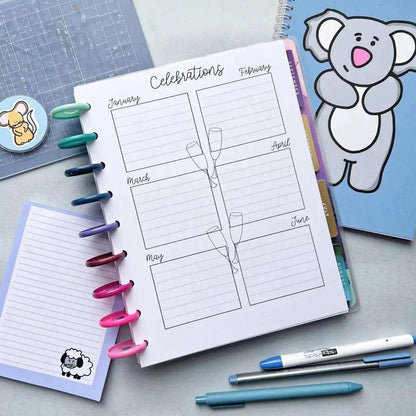 Displayed is the Birthday And Celebration Calendar, opened to the "Celebrations" page featuring monthly boxes and a festive champagne glasses illustration. Adjacent to this birthday planner are a notebook adorned with a koala on the cover, vibrant pens, a blue cutting mat, and sticky notes showcasing delightful sheep and cat designs.