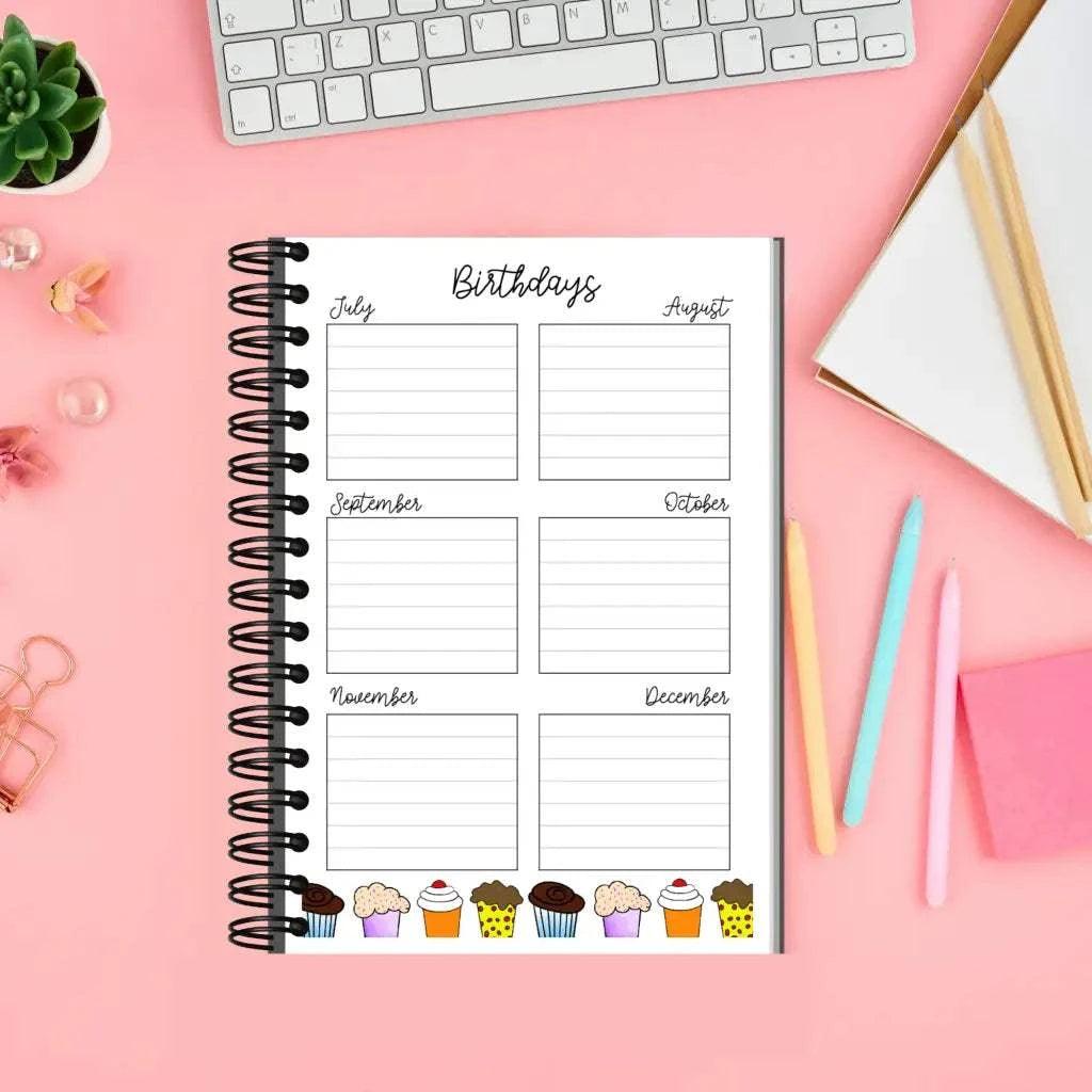 A spiral-bound Birthday And Celebration Calendar lies open on a pink desk featuring sections for July to December, each decorated with cupcake illustrations. Nearby are a keyboard, a plant, a notebook, pens, a pencil, and paper clips—perfectly enhancing the organized workspace for tracking special occasions.