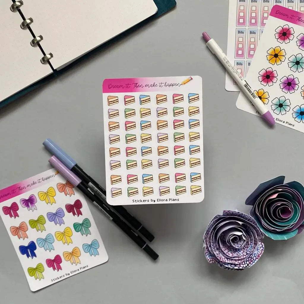 A flat lay photo of a desk with various planning stickers, including a Birthday Cake Planner Stickers with colorful cakes, bows, and flowers. Two purple and black markers and a couple of paper flowers are scattered around. An open notebook is partially visible in the top left corner.