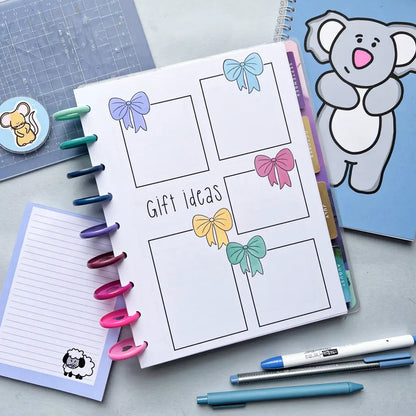 A planner lies open with a page titled "Gift ideas," featuring five sections with bow decorations on each corner indicating different categories. Surrounding the planner are various stationery items, including pens, a notepad, stickers, and another Birthday Calendar with a koala design for efficient birthday planning.