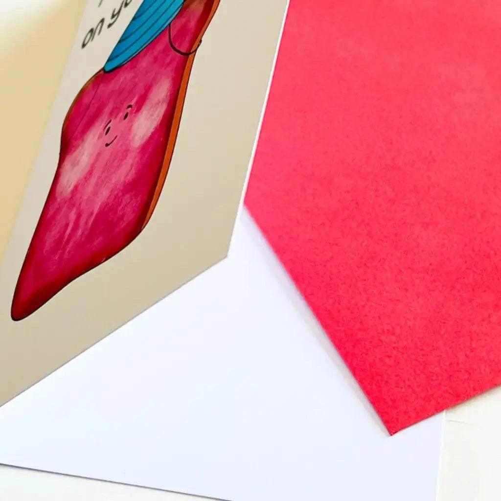 Close-up of the Birthday Toast Birthday Card. The card showcases a pink heart character wearing a blue hat and boasts a metallic appearance. A red envelope is partially visible next to it, making it perfect for someone's special day.