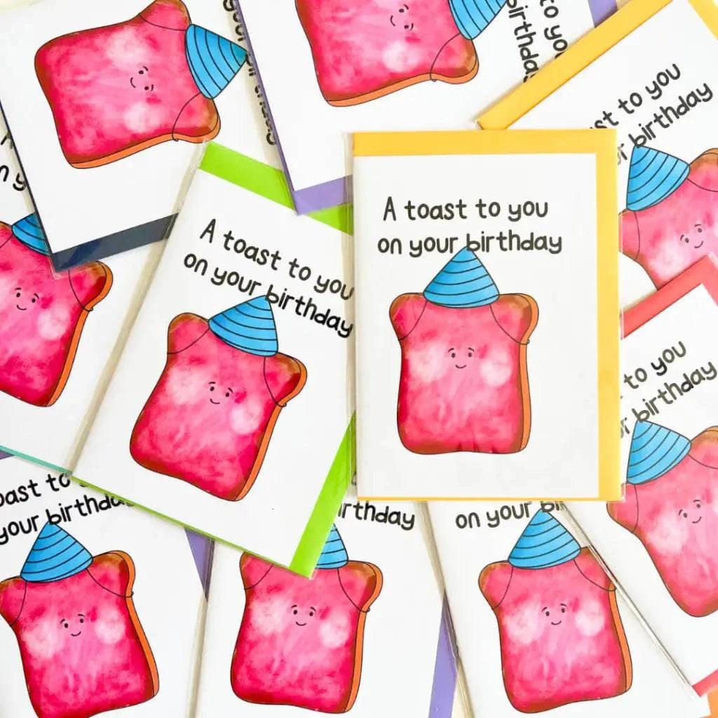 The Birthday Toast Birthday Card is a collection of colorful birthday cards showcasing a cheerful piece of toast donning a blue party hat. The playful illustration style is highlighted with strawberry jam, and the front features the message, "A toast to you on your special day.