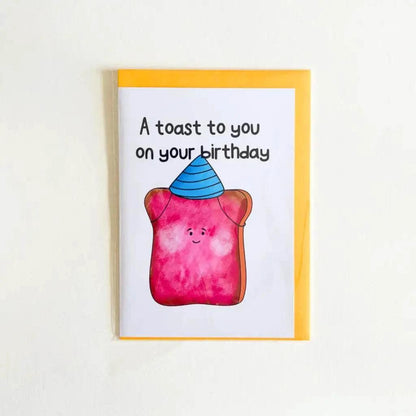 The Birthday Toast Birthday Card showcases a cartoon illustration of a smiling slice of toast donning a blue and white striped party hat, complete with a dollop of strawberry jam. The text above the cheerful design says, "A toast to you on your special day." This delightful card is framed by a yellow border and features a plain white background.