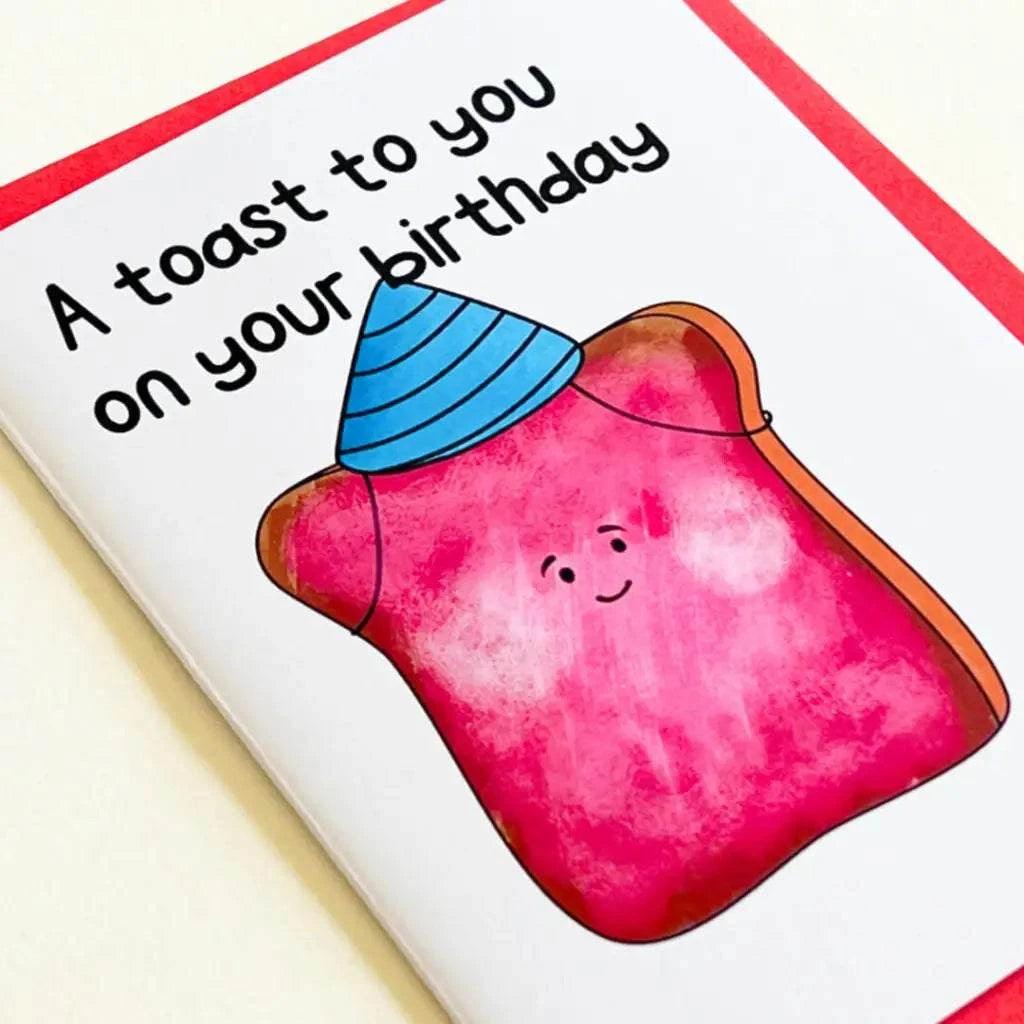 The "Birthday Toast Birthday Card" features an illustration of a smiling piece of toast wearing a blue party hat and topped with strawberry jam. The text on the card reads "A toast to you on your special day." It also has a red border.