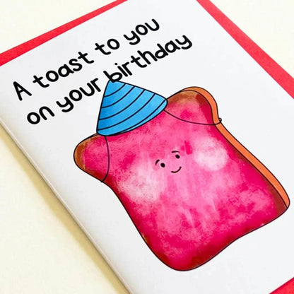 The "Birthday Toast Birthday Card" features an illustration of a smiling piece of toast wearing a blue party hat and topped with strawberry jam. The text on the card reads "A toast to you on your special day." It also has a red border.