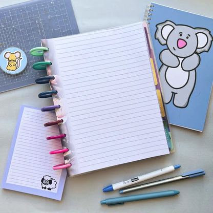 An open notebook with colorful ring binders lays on a table, accompanied by a small lined notepad with a sheep cartoon, a larger koala-themed planner, Blossom Themed Notepaper, a cutting mat with squirrel sticker, and three pens in blue and gray colors.