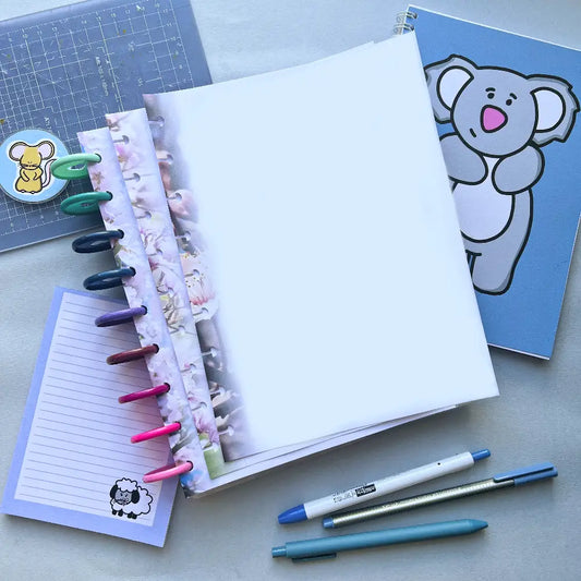 An assortment of stationery is displayed, including a blue notebook with a koala illustration, Blossom Themed Notepaper in a planner, several colored pens clipped to a spiral-bound notebook, a cutting mat, and three pens. The background surface is light gray.