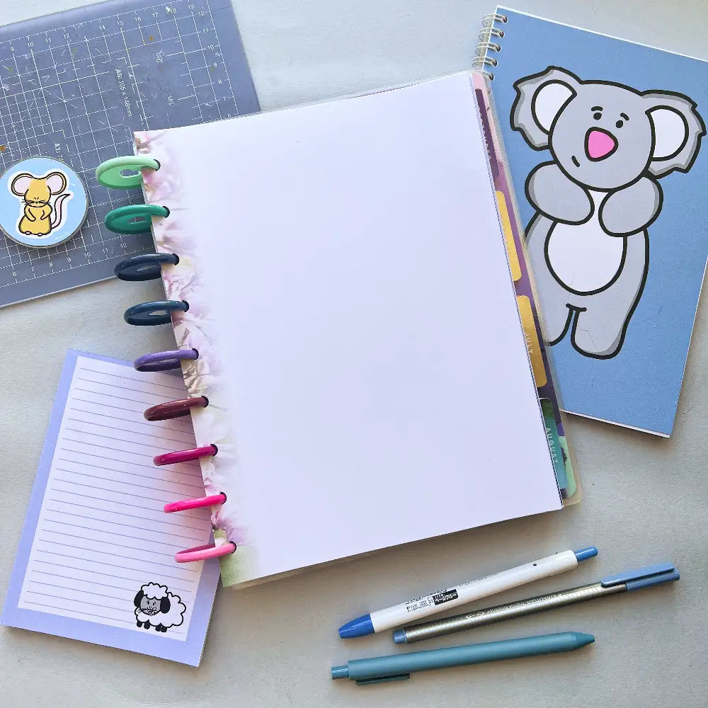 A neatly arranged desk workspace with a blank ring-bound notebook featuring Blossom Themed Notepaper, a small notepad with a sheep design, a koala-themed planner, three pens, and a ruler adorned with a mouse sticker. The stationery items are colorful and organized, ready for use.