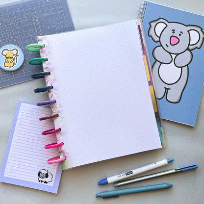 A neatly arranged workspace featuring a dotted spiral-bound notebook with colorful tabs and binder clips. Surrounding it are a blue grid paper pad, a notebook with a cartoon koala cover, a lined notepad with a sheep illustration, three pens, and an elegant planner filled with Blossom Themed Notepaper.