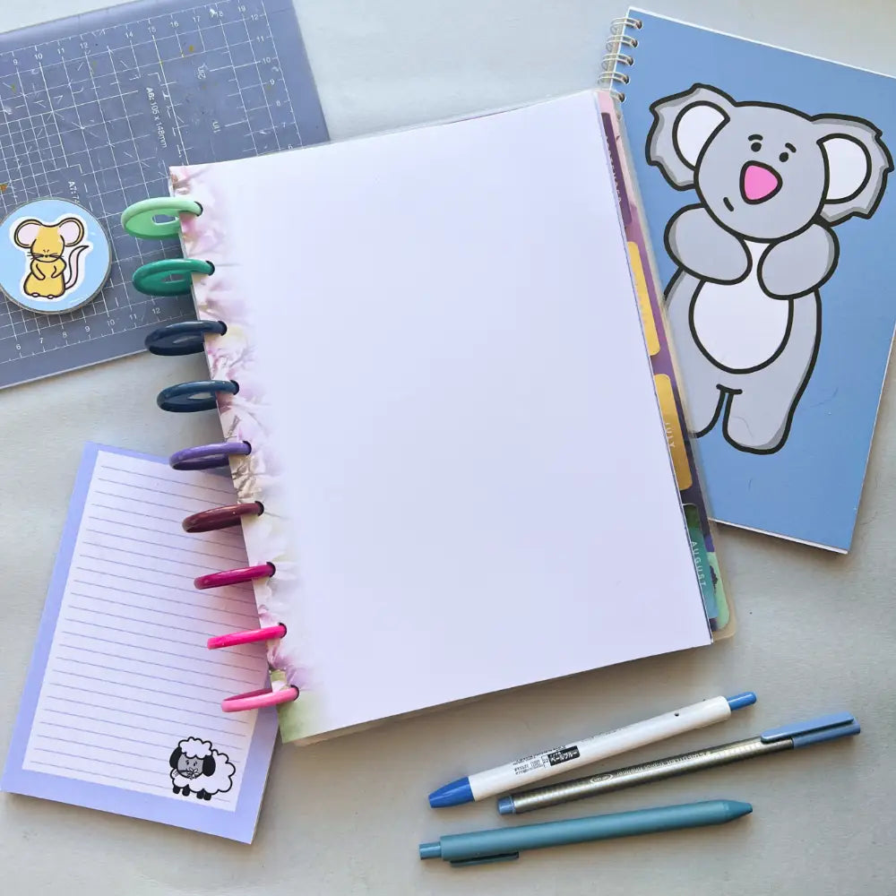 An open notebook with colorful tabs and a blank page is surrounded by a cute koala planner, a lined notepad with a sheep illustration, Blossom Themed Notepaper, a cutting mat, a mouse sticker, a blue pen, a gray pen, and a white and blue pen on a light surface.