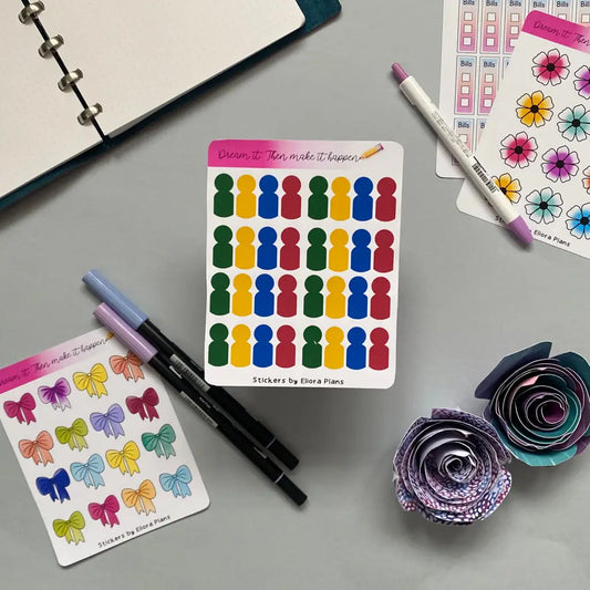 A flat lay of colorful Board Game Players Planner Stickers, pens, and crafted paper flowers on a gray surface. The sticker sheets feature various designs such as silhouettes, flowers, leaves, and board game characters. A notebook is partially visible in the top left corner, perfect for planner decoration enthusiasts.