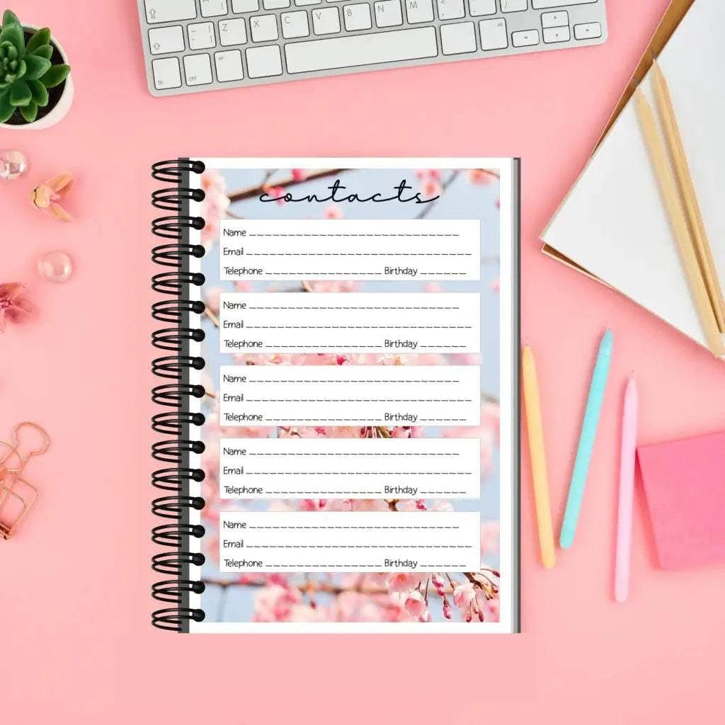 A Blossom Address Book Pages is open on a pink desk, displaying lined entries for Name, Email, Telephone, and Birthday. Surrounding it are a white keyboard, potted succulent, open planner, pastel pens, sticky notes, and a binder clip. The background features pink blossoms.