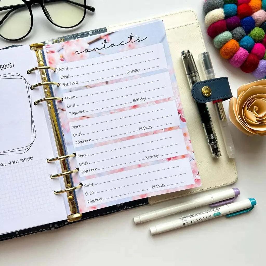 An open planner with a "Contacts" section, providing spaces for name, email, telephone, and birthday. Surrounding items include glasses, colorful felt balls, pens, a paper flower, and two markers. This elegant Cherry Blossom Address Book Pages rests on a white surface.