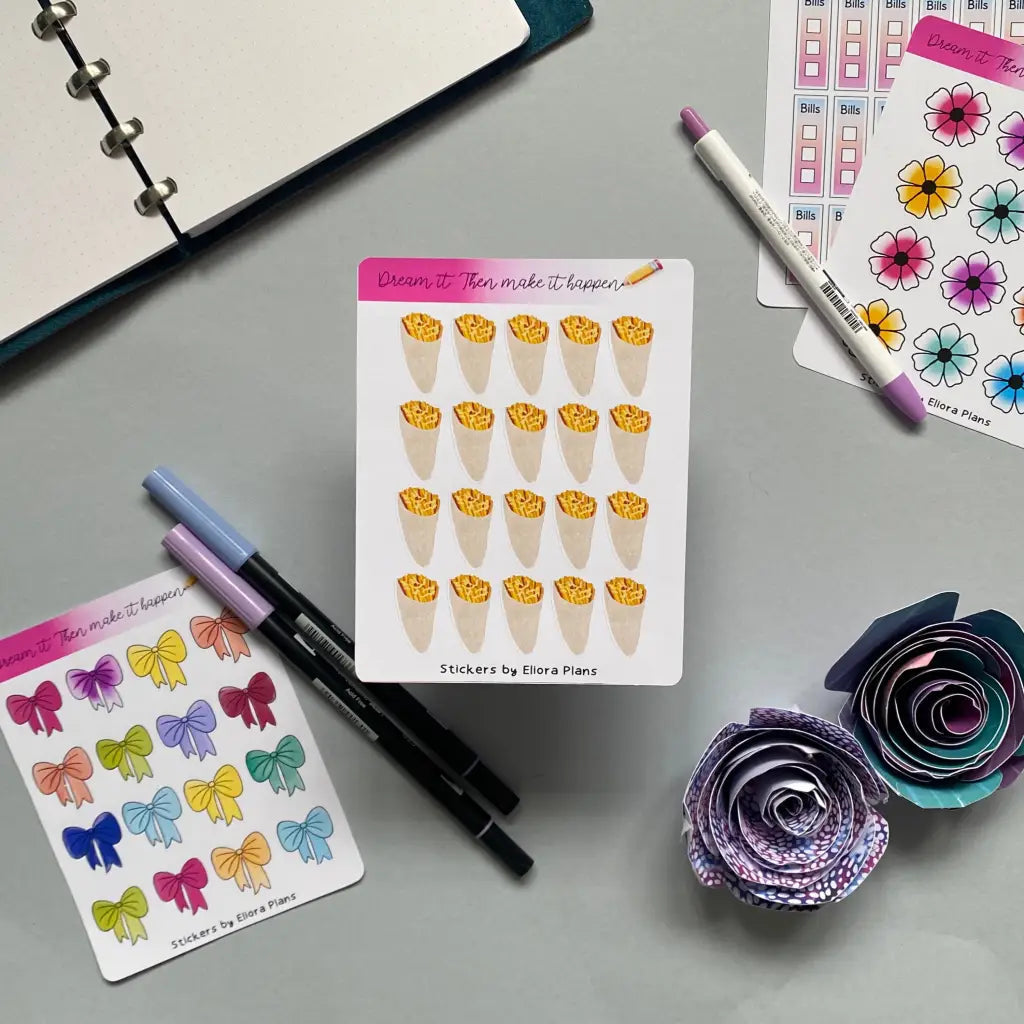 A flat lay on a grey surface showcasing a planner, markers, and floral origami. In the center are three sheets of Chip Cone Planner Stickers: one with tacos, one with colorful bows, and one with flowers. Surrounding the Chip Cone Planner Stickers are pens and a paper flower.