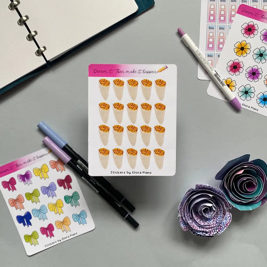 A flat lay of colorful planner stickers, including flower and plant designs, arranged on a gray surface. Nearby are two pens, a purple fine-tipped marker, paper flowers, and an open planner. The decorative stickers, some featuring Chip Cone Planner Stickers themes, have motivational headers and various decorative items.