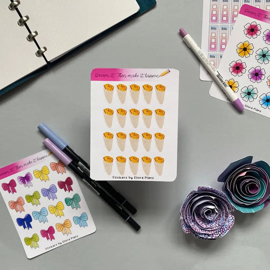 An assortment of colorful stationery items is arranged on a gray surface. There are Chip Cone Planner Stickers, pens, a notebook, and paper flowers. The decorative stickers feature various designs including flowers and bows, and the text "Dream it. Then make it happen.