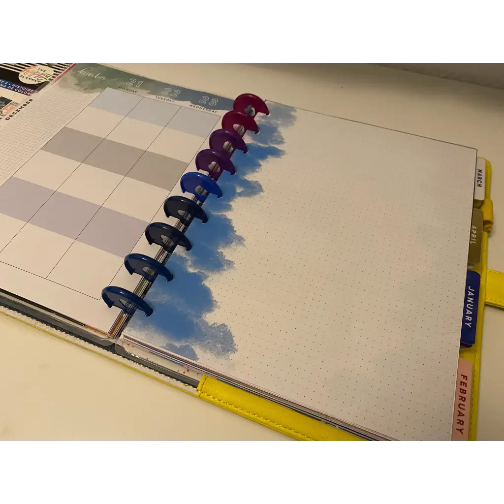 A close-up of an open planner with colorful disc-bound rings on the left edge. The left page displays a grid layout for planning, while the right page is blank with a blue cloud design. Tabs for January and February are visible on the right side, making it perfect for fans of Cloudy Notepaper.