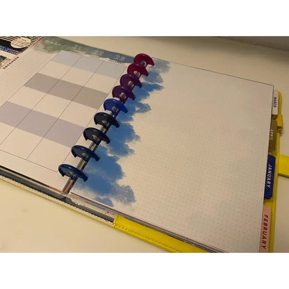 A close-up of an open planner with colorful disc-bound rings on the left edge. The left page displays a grid layout for planning, while the right page is blank with a blue cloud design. Tabs for January and February are visible on the right side, making it perfect for fans of Cloudy Notepaper.