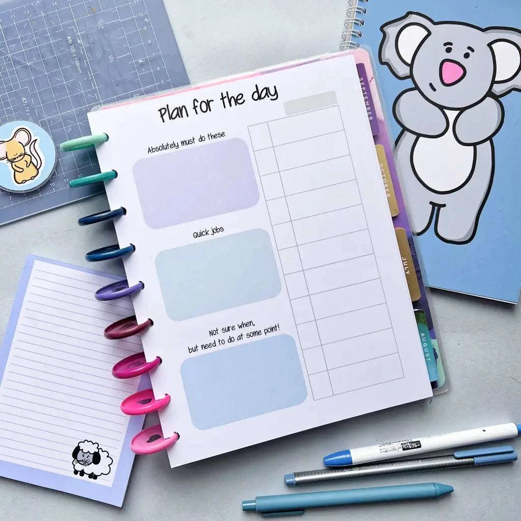 An open Daily Schedule with sections for "Absolutely must do these," "Quick jobs," and "Not sure when, but need to do at some point" lies on a desk, serving as an excellent tool for task management. Nearby are a blue notebook with a cartoon koala on the cover, a small notebook with a sheep sticker, and several pens.