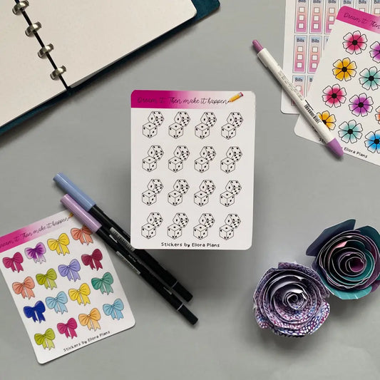 Top view of a desk with various colorful stickers, markers, pens, and paper flowers. The Dice Planner Stickers include illustrated D6 dice, checkboxes, and flowers. An open planner is partially visible in the top left corner featuring a themed planner page. The scene is well-organized and colorful.