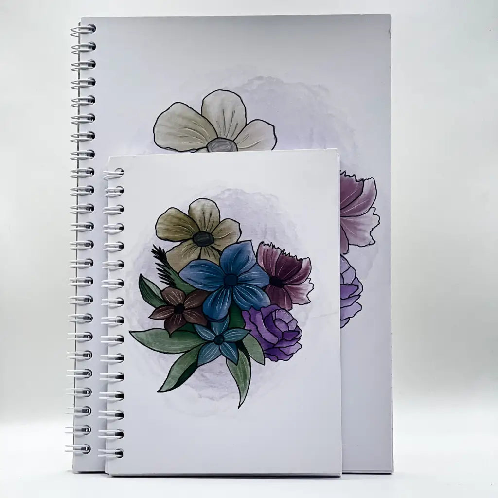 Two spiral-bound Faded Floral Notebooks are displayed against a plain background. The larger notebook is behind the smaller one. Both have white covers featuring a faded floral design with blue, purple, and beige flowers with green leaves.