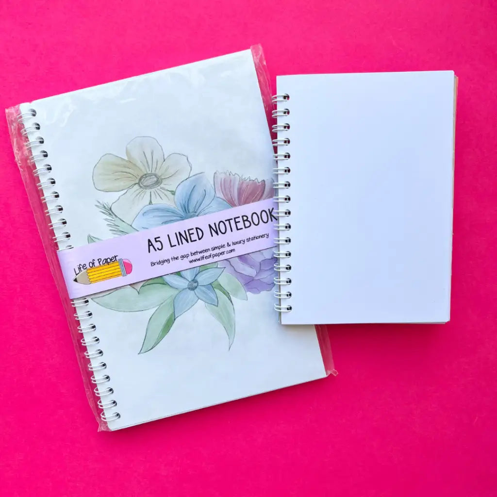 Two spiral-bound A5 notebooks are shown against a bright pink background. The notebook on the left, a vintage-themed with faded floral design, contrasts elegantly with the one on the right, which has a plain white cover. Both high-quality notebooks are labeled "Faded Floral Notebook" and wrapped in plastic.