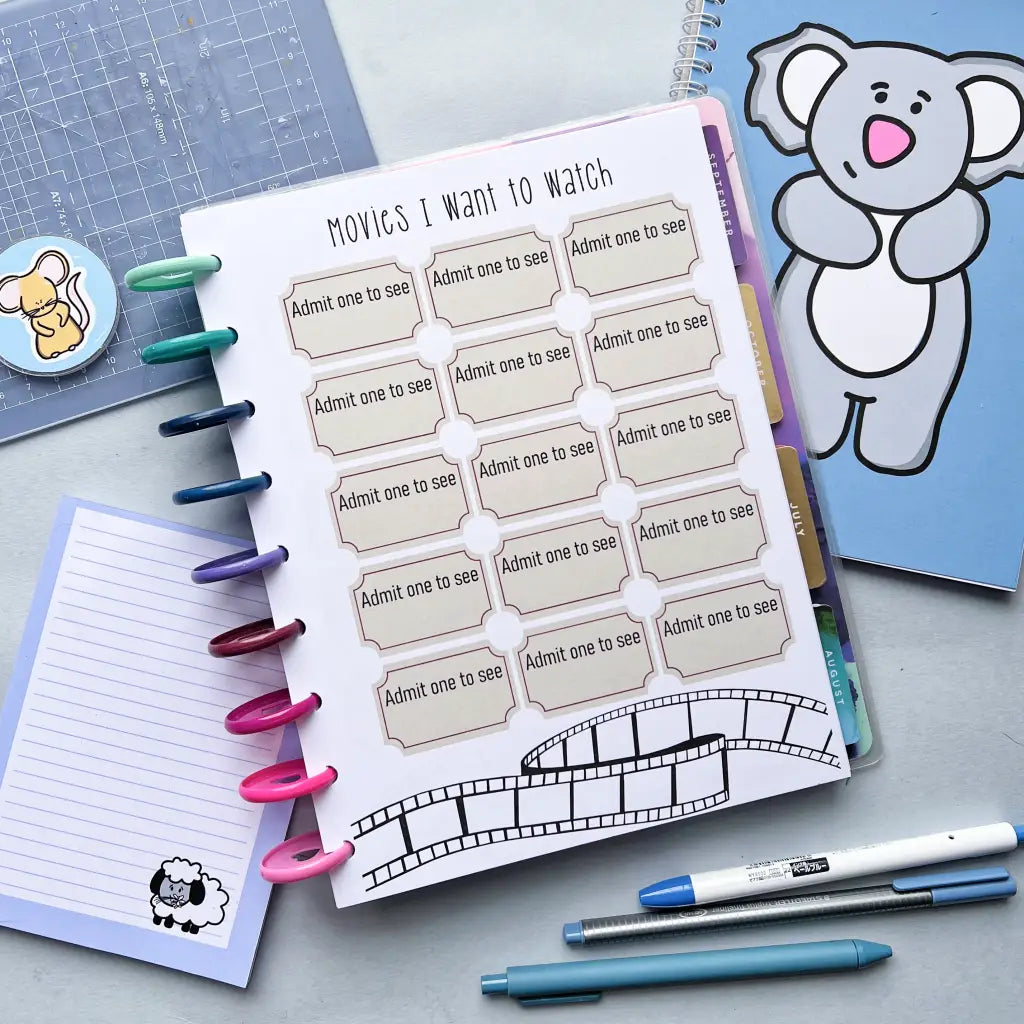 A planner page labeled "Film Tracker" features blank tickets with "Admit one to see" text, perfect for your movie tracker. The page is decorated with film strip graphics. Surrounding the planner are colorful pens, a blue notebook with a koala, sticky notes, and a pen with a sheep design.