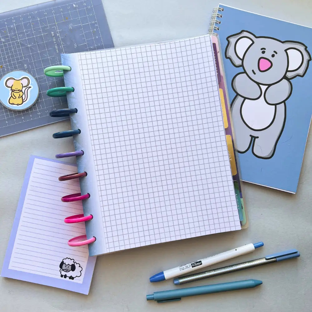 A flat lay photo of stationery items including a Forest Themed Notepaper with multicolored tabs and spiral binding, a journal with a cartoon koala on the cover, a small lined notepad with a sheep illustration, two pens, and a pencil, all arranged on a light-colored surface.