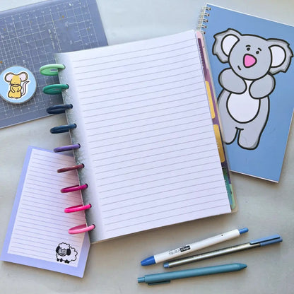 An open notebook with colorful tabbed rings is placed on a desk. Next to it are a blue planner with a koala cover, a small lined notepad with a sheep illustration, and three pens. A Forest Themed Notepaper is also visible on a plastic sheet at the side.