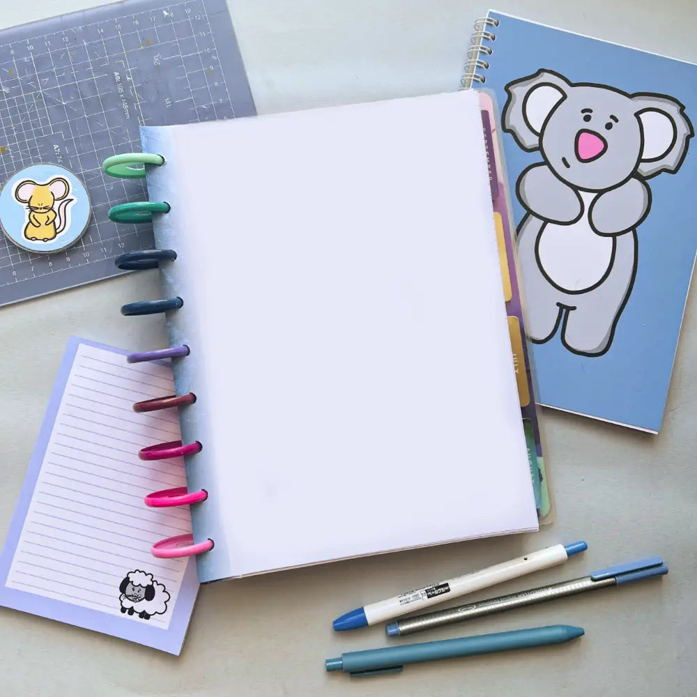 A flat lay of stationery items including a Forest Themed Notepaper, a gray journal with a koala illustration, a small lined notepad with a sheep illustration, a cutting mat with a mouse sticker, three pens, and a crochet hook on a light surface.