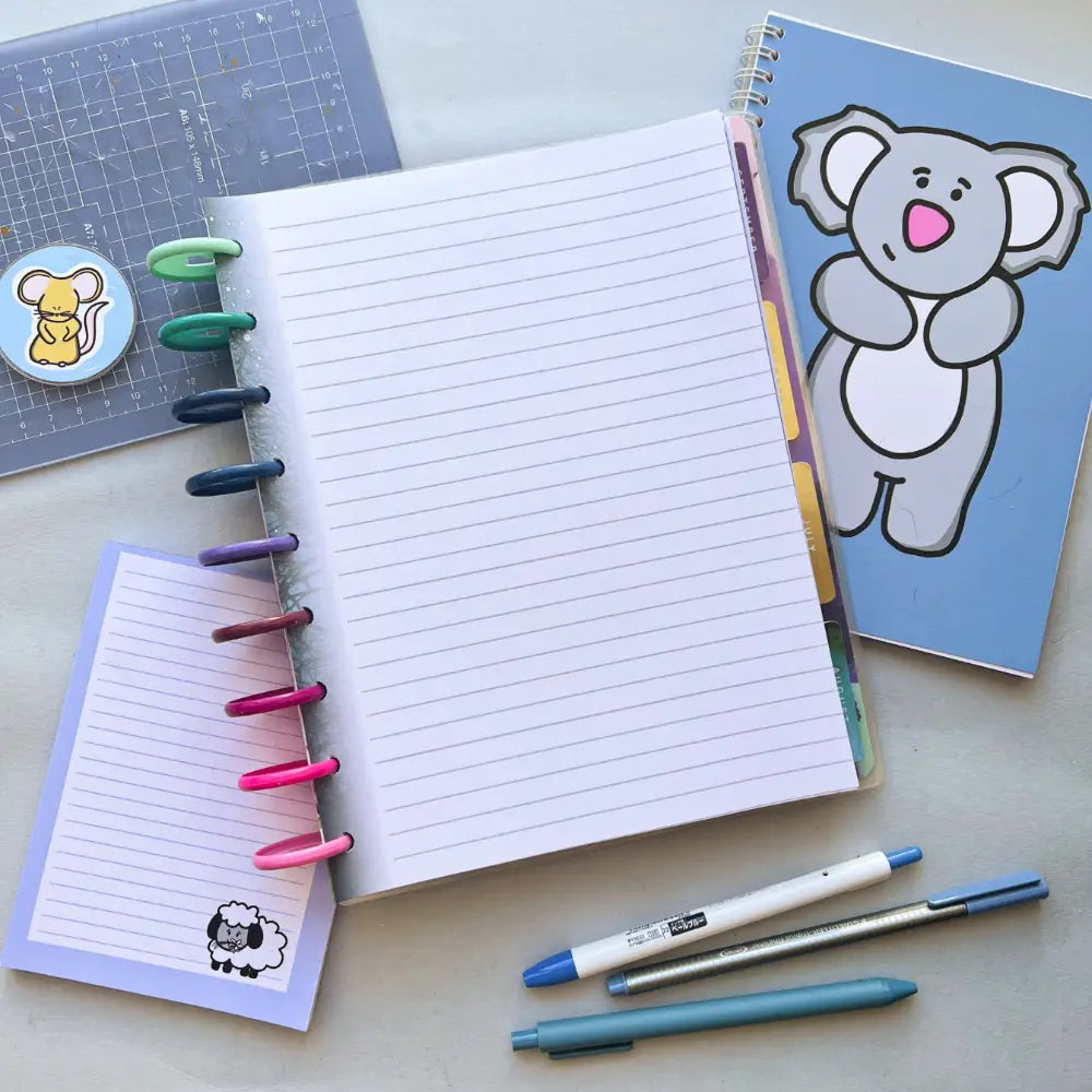 An open notebook with colored tabs and lined pages lies on a desk. Nearby, there's Forest Themed Notepaper, a pen, and a blue journal with a koala drawing. A cutting mat with a mouse sticker is in the background, and three pens are placed beside the notebooks and planner.