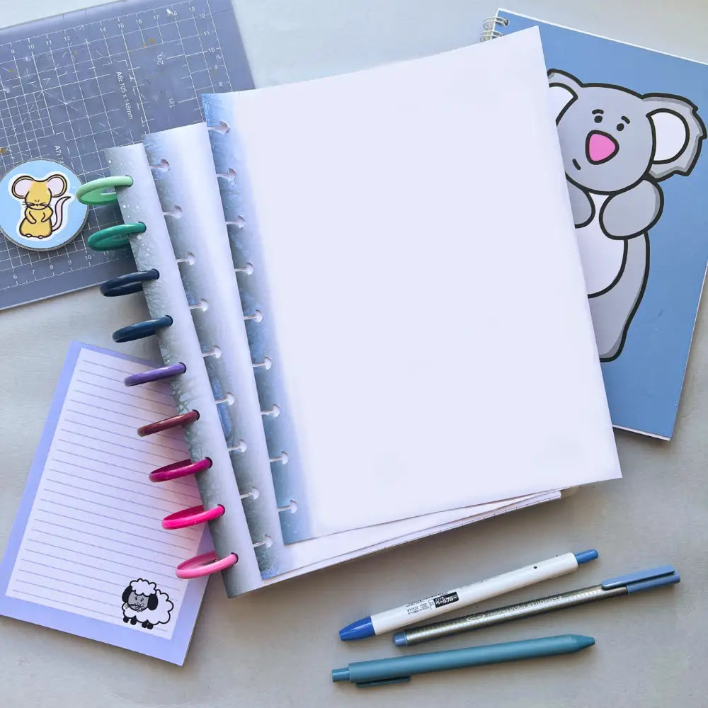 An assortment of stationery items is spread out on a table: a blank notebook with colorful rings, a blue notebook with a koala illustration, Forest Themed Notepaper, two pens, and a sticker of a squirrel. A planner filled with ample filler paper and a blue cutting mat are also visible.